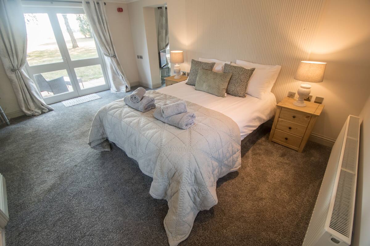 Habitación de hotel con una cama grande, una mesita de noche, una lámpara y vistas al exterior a través de una puerta corredera.