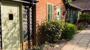 Une maison en briques avec une porte et des volets verts, une petite fenêtre et un jardin agrémenté de fleurs et d’arbustes.
