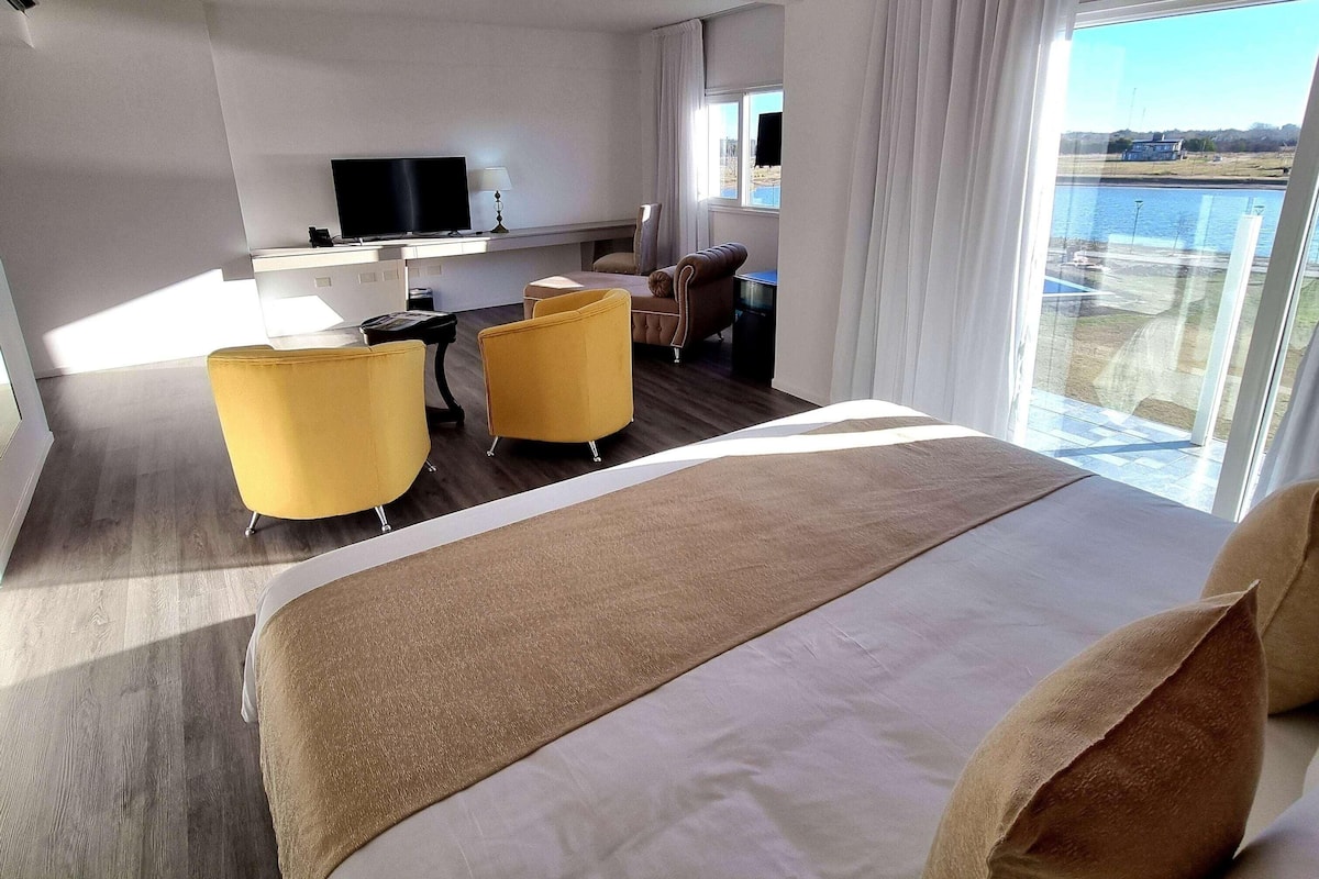 Hotellihuone, jossa on suuri sänky, kaksi keltaista nojatuolia, työpöytä, jossa on televisio, ja näkymä vesistöön.