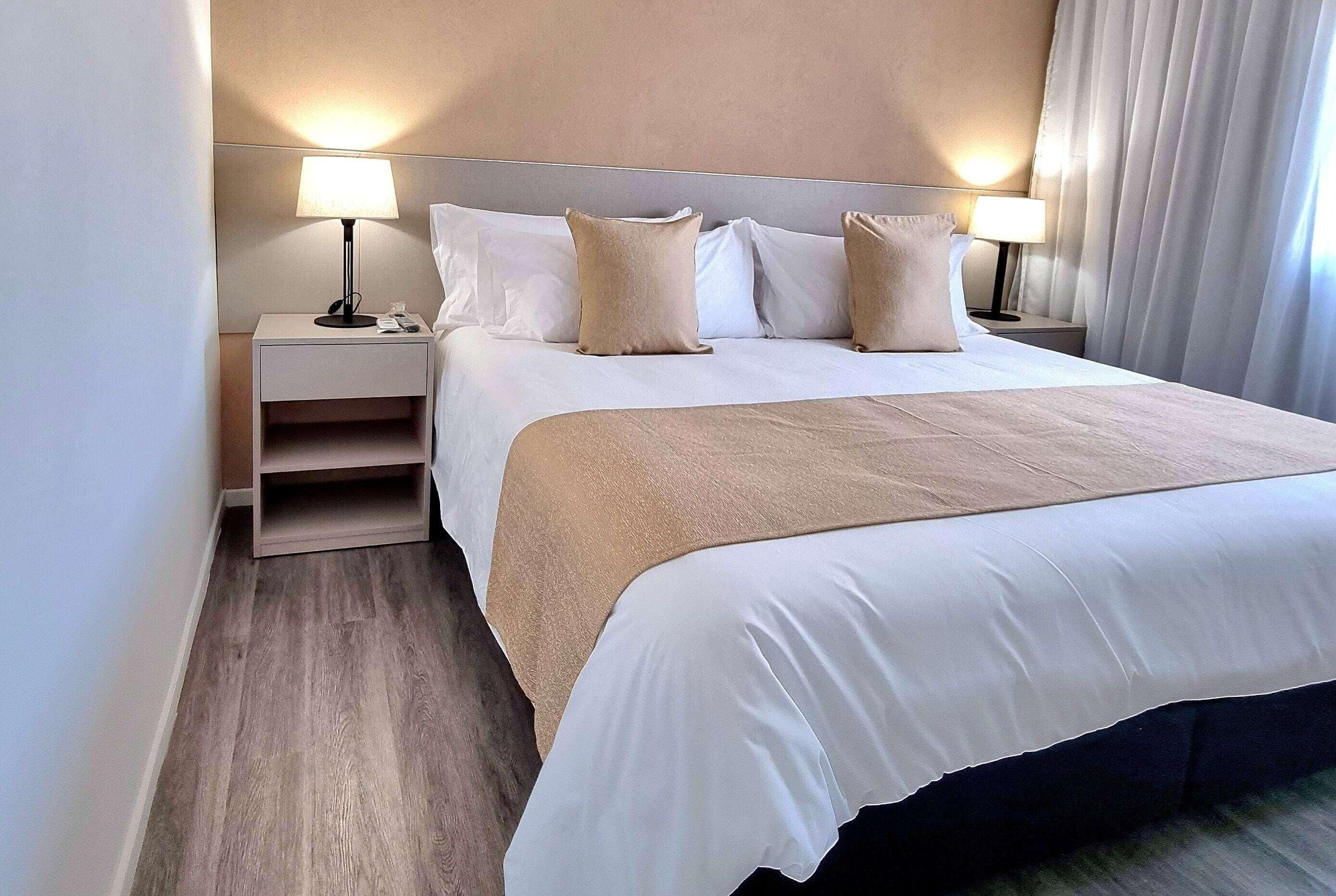A neatly made bed with white linens and a beige throw, flanked by two bedside tables with lamps, in a room with wooden flooring and neutral-toned walls.
