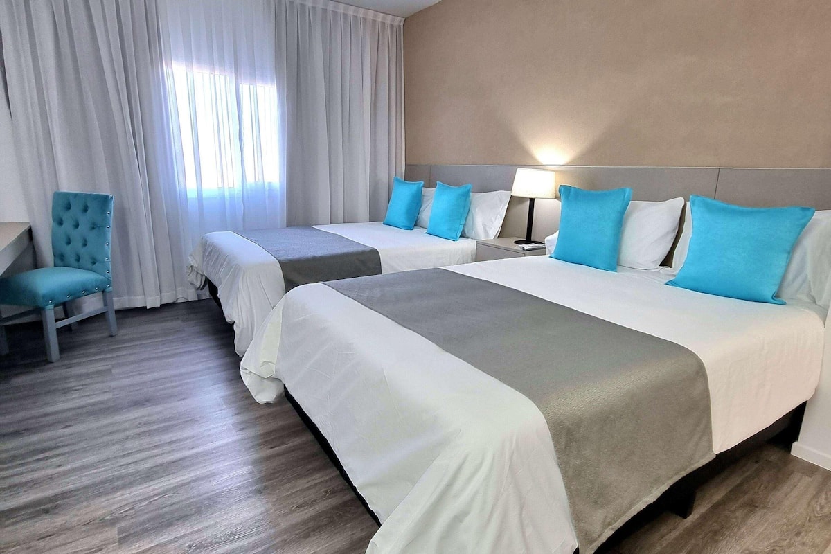 Hotellihuone, jossa on kaksi sänkyä, tuoli, yöpöytä ja lamppu.