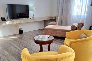 A hotel room with a flat-screen TV, a small table with a magazine, a yellow armchair, a pink sofa, and a lamp.