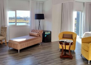 A room with a single bed, a chair, a table, a lamp, and large windows.