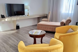 A hotel room with a flat-screen TV, a small table with a magazine, a yellow armchair, a pink sofa, and a lamp.