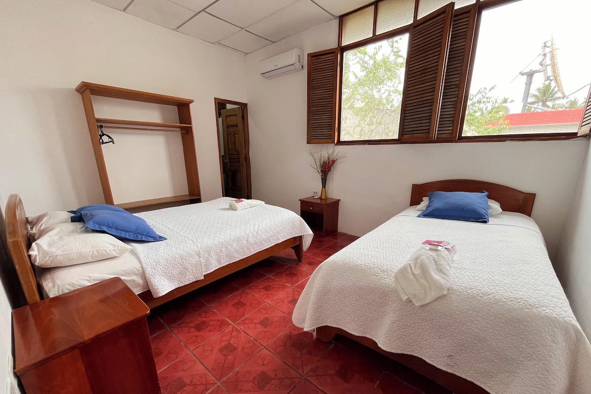 Two single beds with wooden headboards, a wooden shelf unit, a small wooden nightstand with a vase, and a window with wooden shutters.