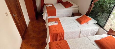 A hotel room with three beds, each with white bedding and orange accents. There is a wooden door and a window with curtains.