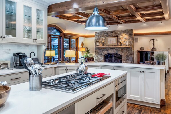 A kitchen with a large island, white cabinetry, a stone fireplace, and a hanging pendant light.