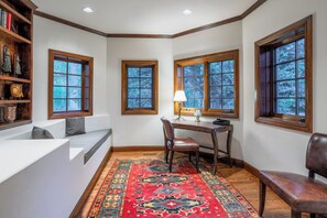 Habitación de esquina con un banco empotrado, una mesa con una silla y un ventanal con vista a árboles.