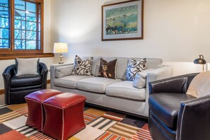 Una sala de estar con un sofá gris, dos sillones, un reposapiés rojo y una alfombra estampada.