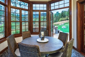 A round table with wicker chairs and a candle centerpiece, surrounded by large windows with a view of a green landscape.