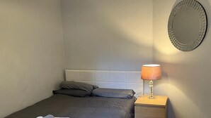 Una cama bien tendida con una colcha oscura y almohadas, una mesita de noche con una lámpara, y un espejo montado en la pared.