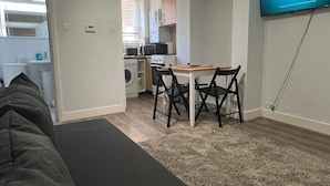 Un espacio habitable compacto con sofá, una mesa de comedor con sillas, televisor montado en la pared y una cocina visible a través de una puerta abierta.