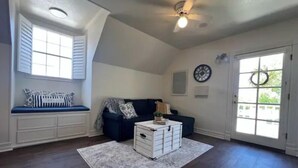 A spacious room with a high ceiling, a built-in bench with storage, a dark-colored sofa, a white chest of drawers, and a large window with shutters. - Sunsets & Shows! Gorgeous, Private 1 BR Listing! (Mesa)