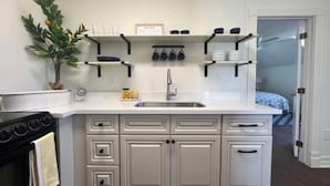 A compact kitchen with white cabinetry, a built-in oven, and a sink. There are shelves above the sink holding dishes and a small potted plant. - Sunsets & Shows! Gorgeous, Private 1 BR Listing! (Mesa)