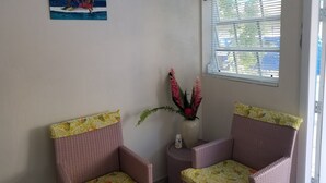 Ein Zimmer mit zwei Korbstühlen, einer Wanduhr, einem Fenster mit Jalousien und einem Bild von zwei Personen am Meer.