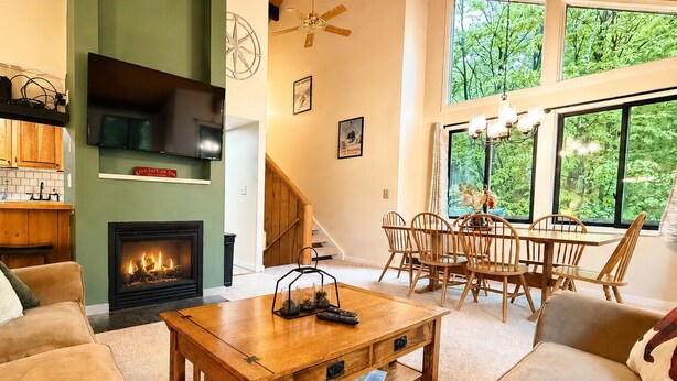 Un salon doté d’une cheminée, de meubles en bois, d’un coin repas et de grandes fenêtres offrant une vue sur la verdure.
