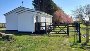 A small, single-story white building with a sloped roof, a wooden gate, and a grassy area in front. - The Paddocks -  a bungalow situated in a rural village surrounded by fields and horses (Frettenham)
