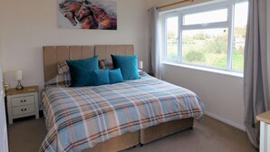 A bedroom with a bed, a nightstand, a lamp, a window with curtains, and a painting on the wall. - The Paddocks -  a bungalow situated in a rural village surrounded by fields and horses (Frettenham)