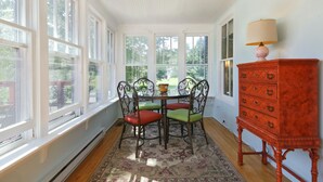 Une véranda avec une table et des chaises, de grandes fenêtres, une commode en bois et une lampe.