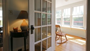 Une pièce avec un plancher en bois, un fauteuil à bascule, une lampe sur une table et une grande fenêtre donnant sur des arbres.
