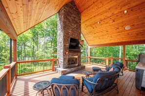 A covered deck with a stone fireplace, outdoor seating, and a view of a forest. - Signature Mountain Lodge w/ Hot Tub & Views (McHenry)
