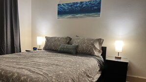 A neatly made bed with a patterned bedspread, two bedside lamps, a clock, and a painting of the ocean on the wall. - #1* 5min walk to the Beach, Amazing Virginia Pro Host. (Norfolk)