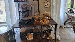 Una cafetería con una máquina Keurig, un cartel que dice "Café primero, ustedes después", y diversos artículos relacionados con café sobre una mesa de madera.
