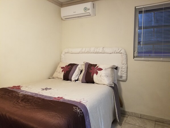 A bedroom with a bed, pillows, a window with blinds, and an air conditioning unit on the wall. - The Perfect Island Getaway #4 (Nassau)