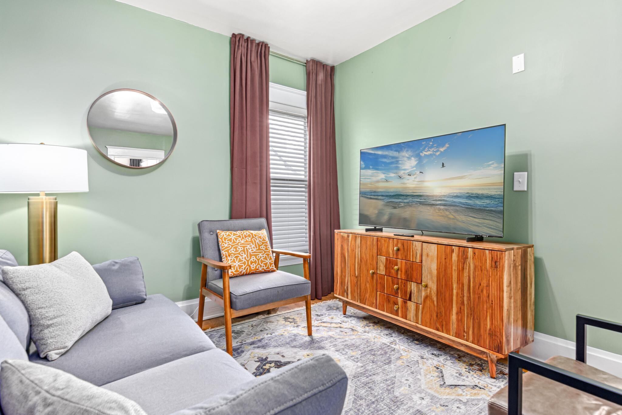 A living room with a grey sofa, a wooden cabinet, a round mirror, and a television on a wall.
