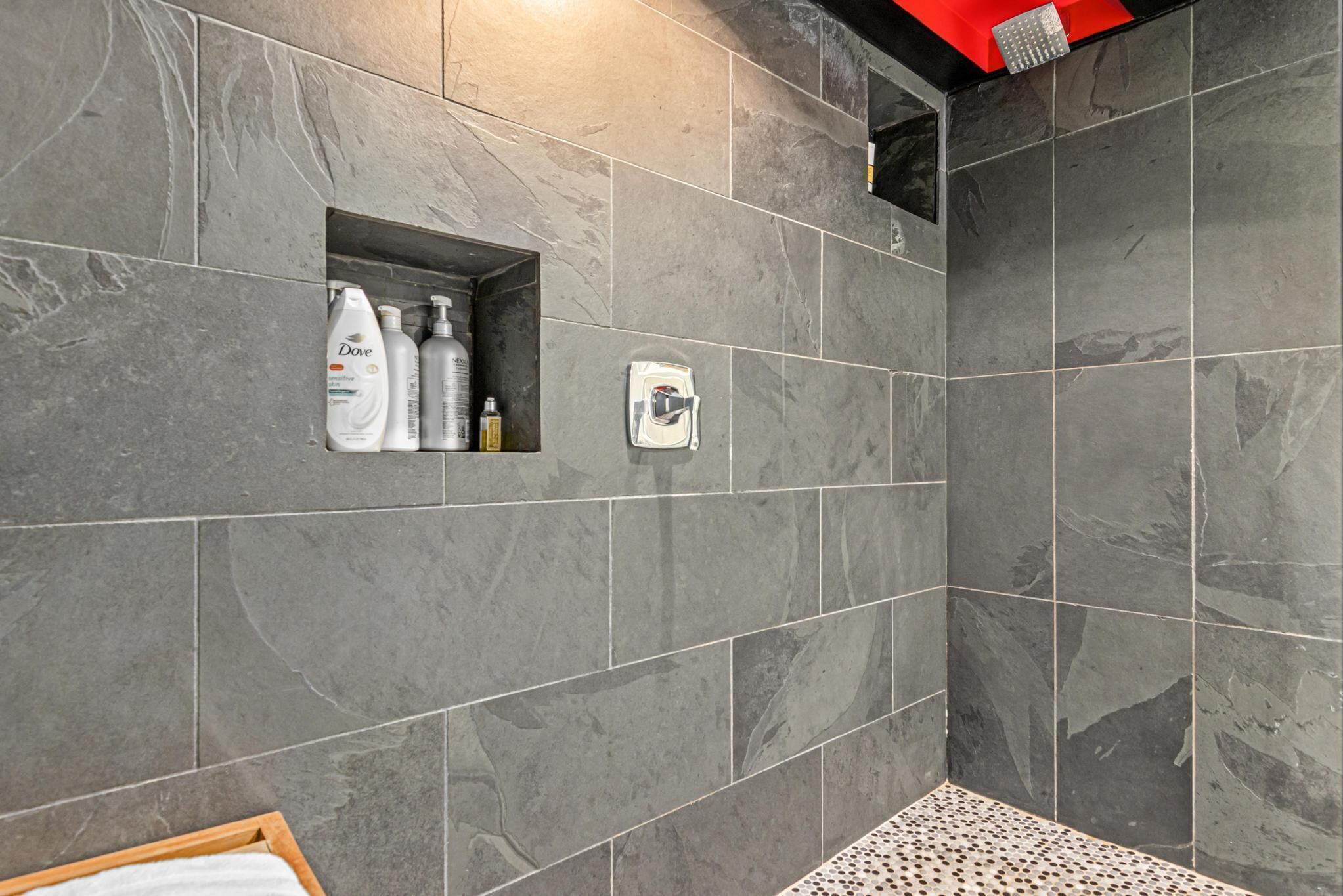 Un baño con baldosas de mármol gris, zona de ducha y estante con artículos de aseo.