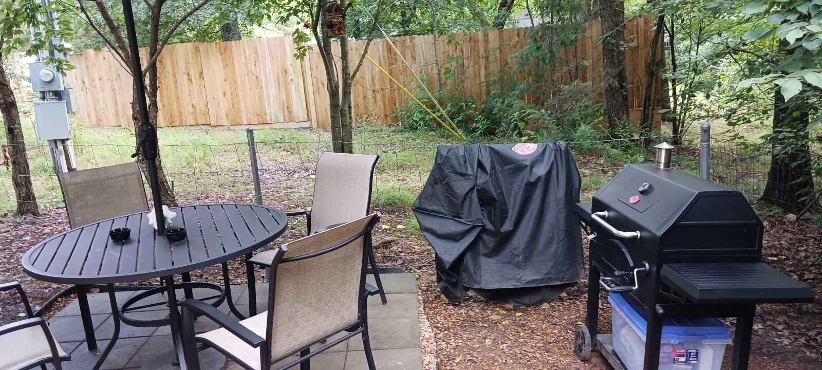 A backyard with a round table and chairs, a covered grill, and a wooden fence.