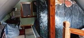 Ein Zimmer mit schrägverdeckter Decke, einem Bett, einem Schreibtisch mit Computer, einem Fenster mit Vorhang und einer Wand mit Waldgemälde.