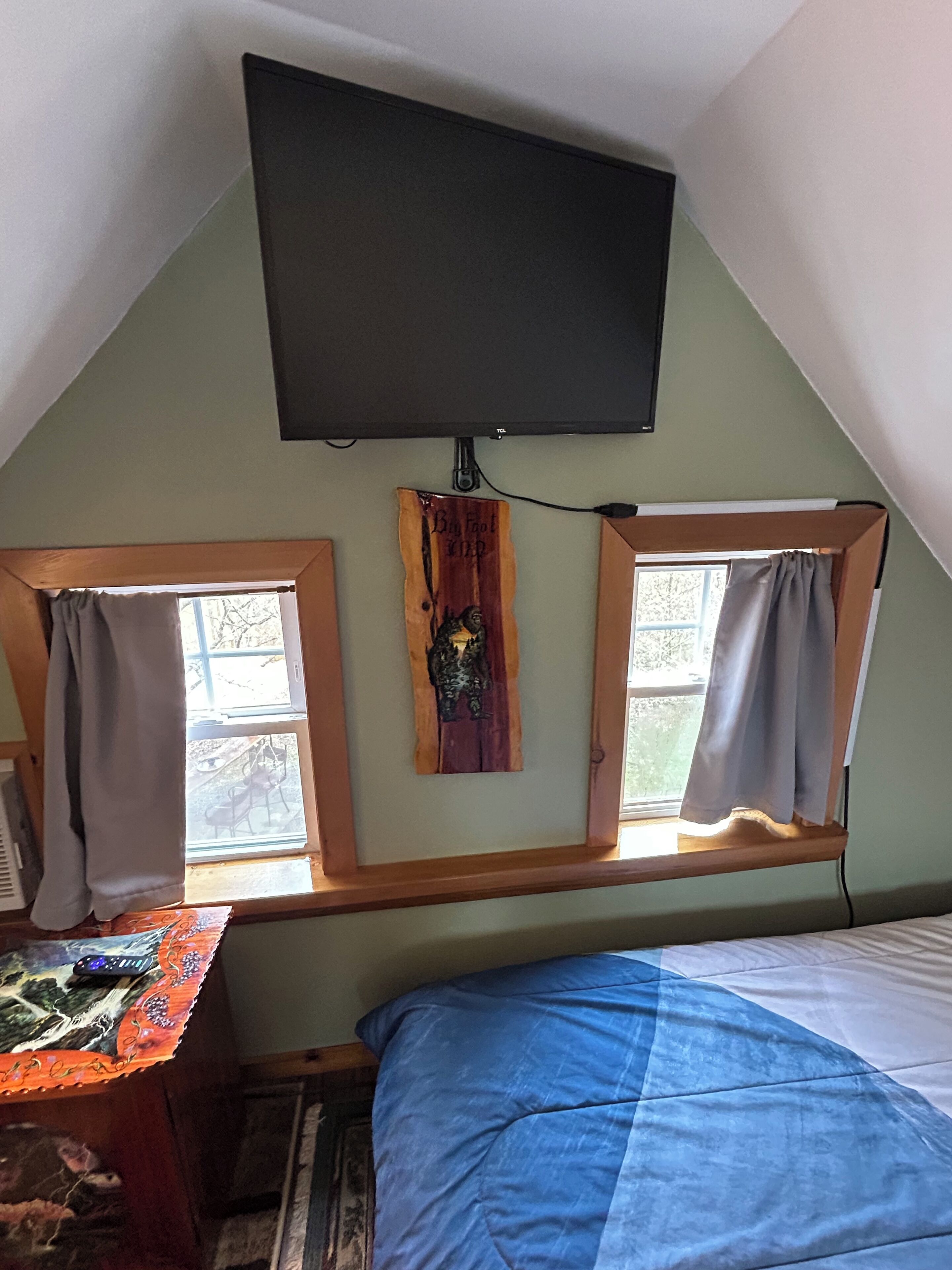 A bedroom with a bed, a TV mounted on the wall, and two windows with curtains.