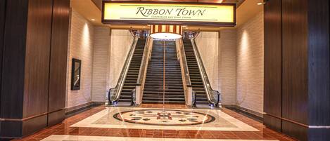 A hotel lobby with a central escalator, a large illuminated sign reading "Ribbon Town Conference and Event Center," and a patterned floor design.