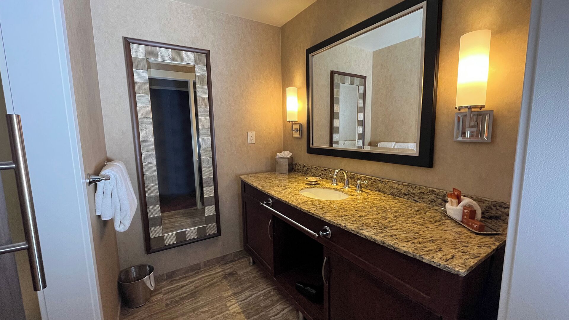 A bathroom with a marble sink, a large mirror, a towel rack, and a trash can.