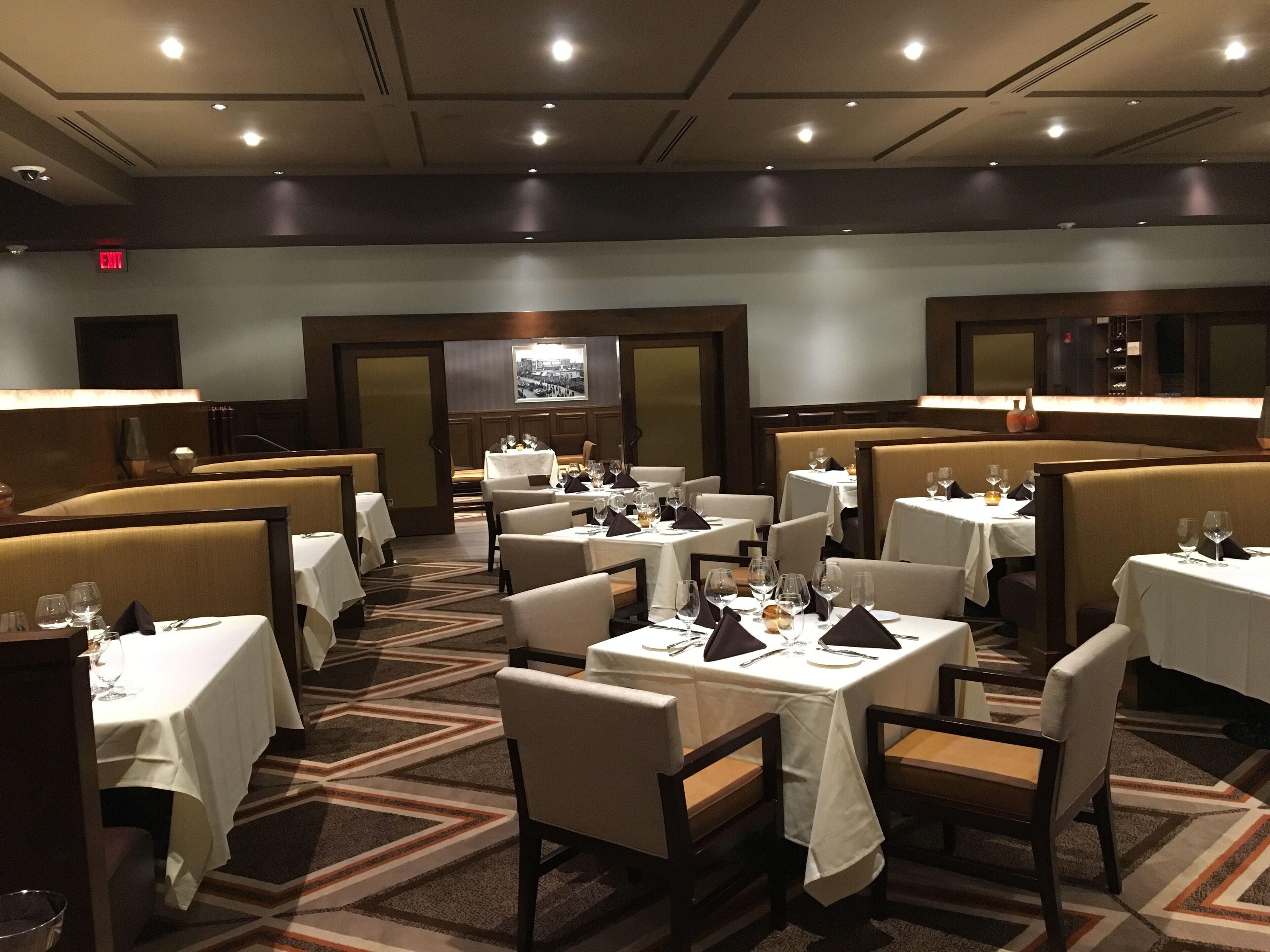 A restaurant with multiple tables and booths, each with white tablecloths and set with wine glasses and cutlery.