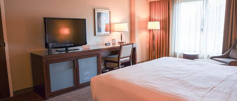 Habitación de hotel con cama, escritorio con televisor, silla, lámpara y ventana con cortinas.