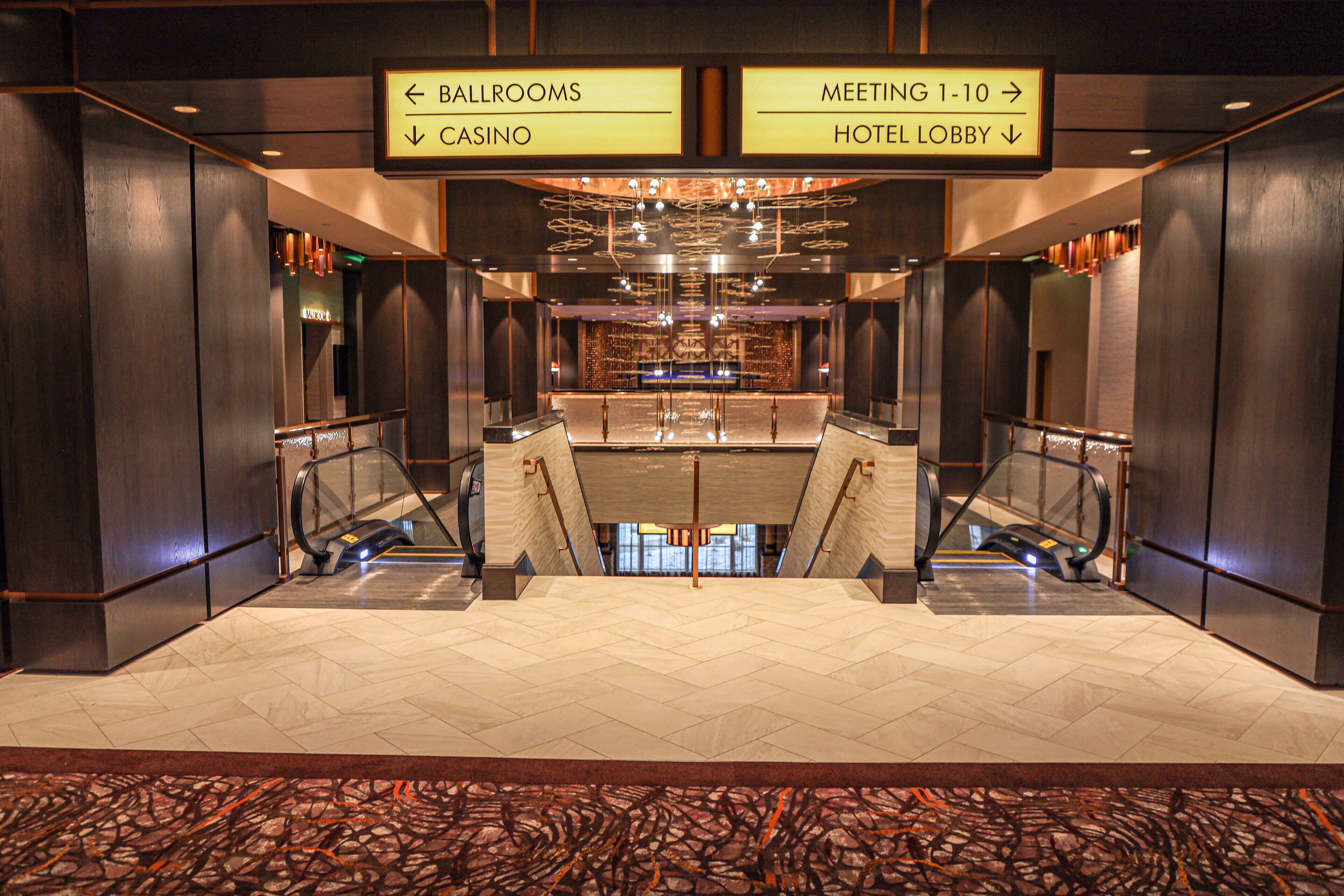 A hotel lobby with an escalator, a reception desk, and directional signs for the ballrooms, casino, meeting rooms, and hotel lobby.