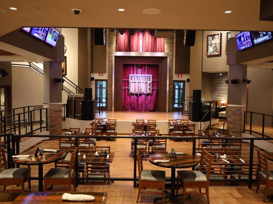 A restaurant with a stage and curtains in the center, wooden tables and chairs, and multiple televisions on the walls.