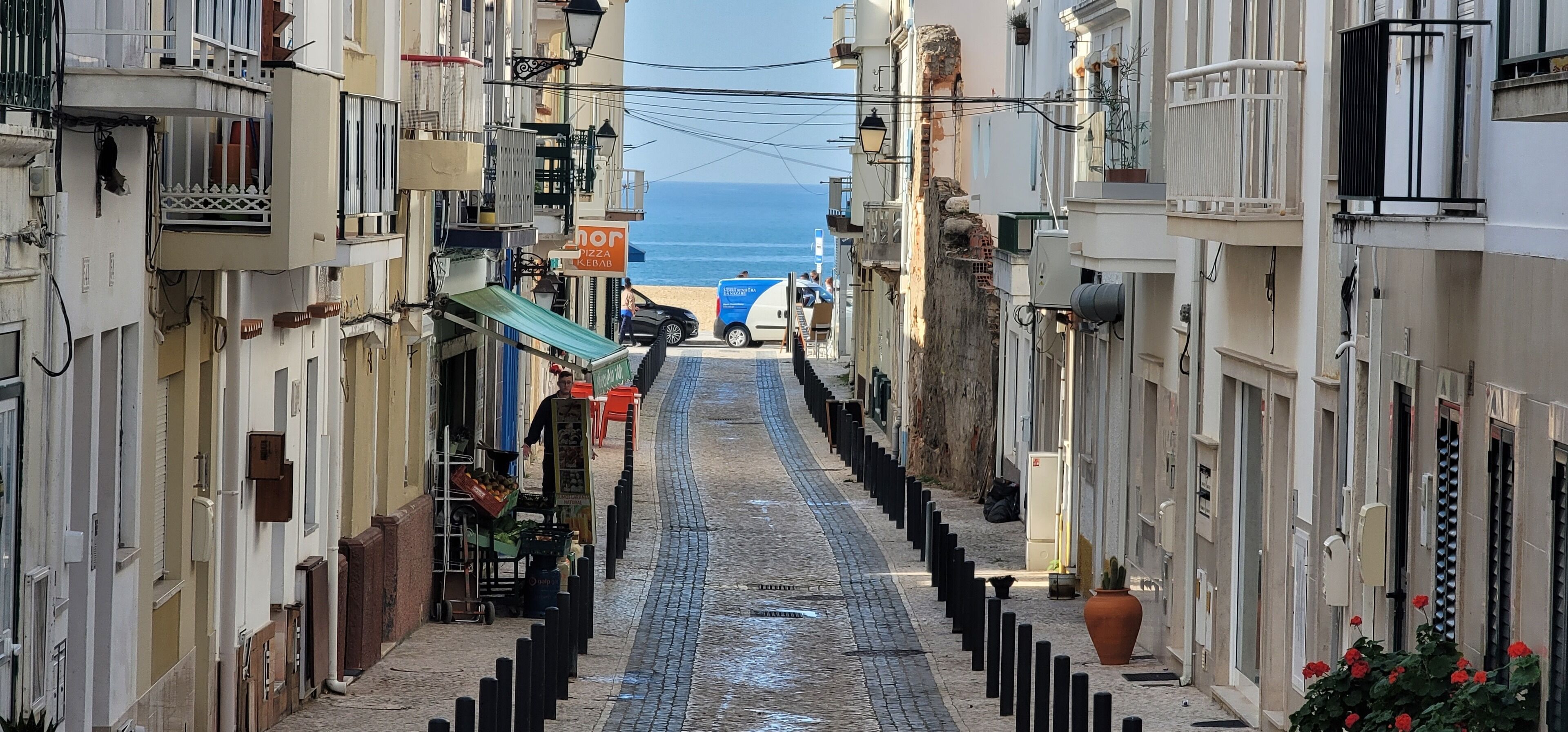 Eine enge Straße, gesäumt von Gebäuden mit Balkonen, mit einem Strand in der Ferne und einem geparkten Wagen.