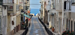 Une rue étroite bordée d’immeubles dotés de balcons, une plage visible au loin et un fourgon garé.