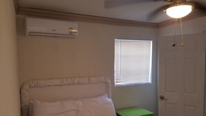 Une chambre à coucher comprenant un lit, un appareil de climatisation, une fenêtre avec des stores, une table de chevet verte et une porte.