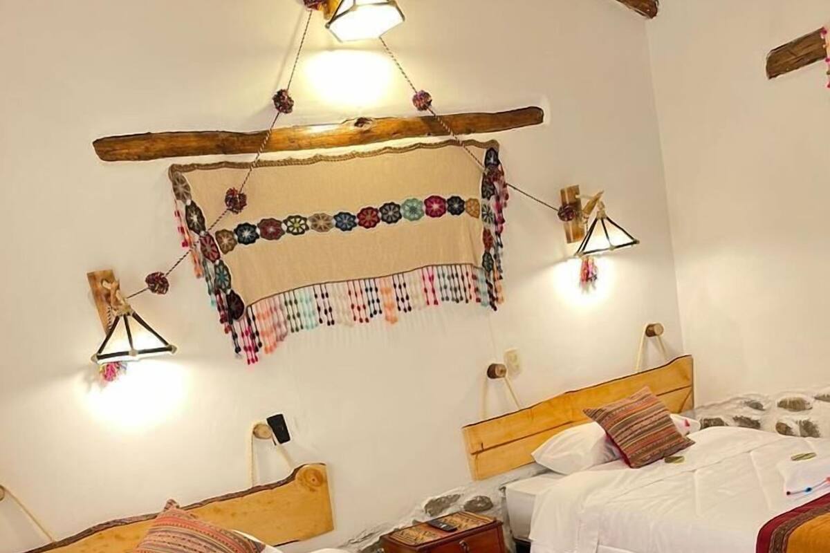 Una habitación con dos camas de madera, cada una con cabecero, ropa de cama blanca y flecos coloridos. Hay luces fijadas a la pared y un adorno colgante sobre las camas.