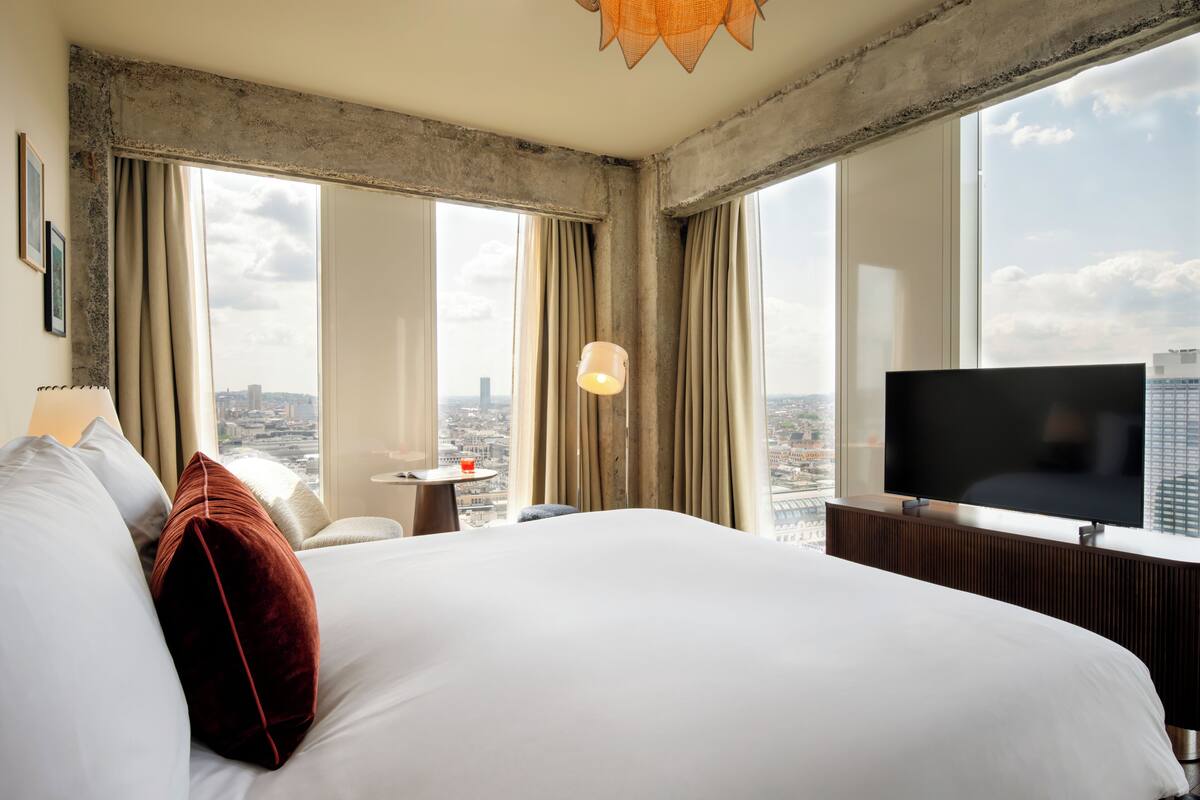 Quarto de hotel com uma cama grande, televisão, mesinha e vista para a cidade através de amplas janelas.