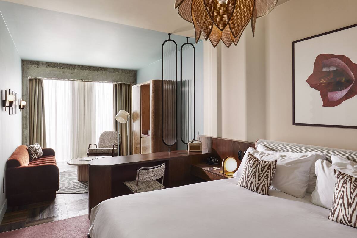 Quarto de hotel com uma cama grande, uma escrivaninha, uma cadeira, um sofá e uma janela ampla com cortinas.