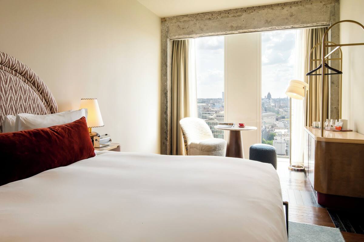 Quarto de hotel com uma cama grande, uma cadeira, uma mesinha e vista da cidade por uma janela ampla.