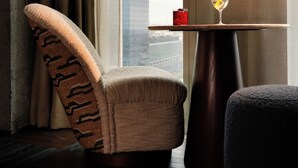 A modern room with a view of a cityscape, featuring a cushioned armchair, a small round table with a drink, and a dark ottoman.