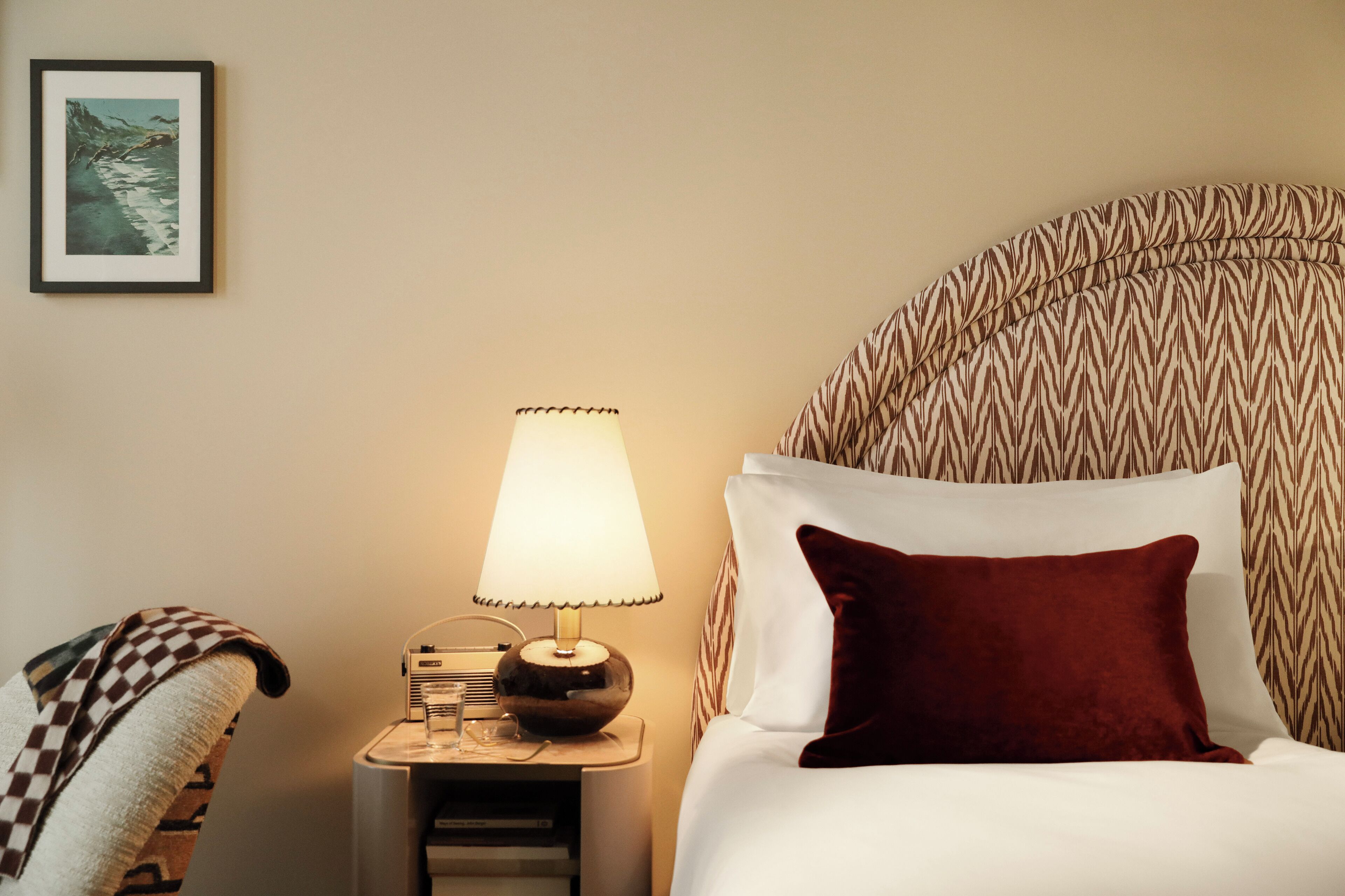 Una cama bien hecha con un almohadón rojo, una lámpara de noche y un cuadro en la pared.