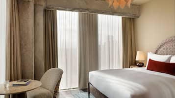 A hotel room with a large bed, a round table, a chair, and a view of the city through large windows.