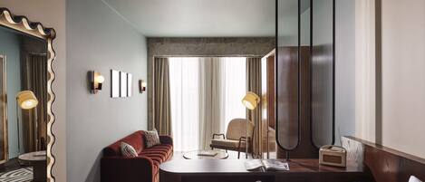 Une chambre d’hôtel moderne avec une table à manger ronde, un canapé, un fauteuil, un grand miroir et une vue sur l’extérieur.